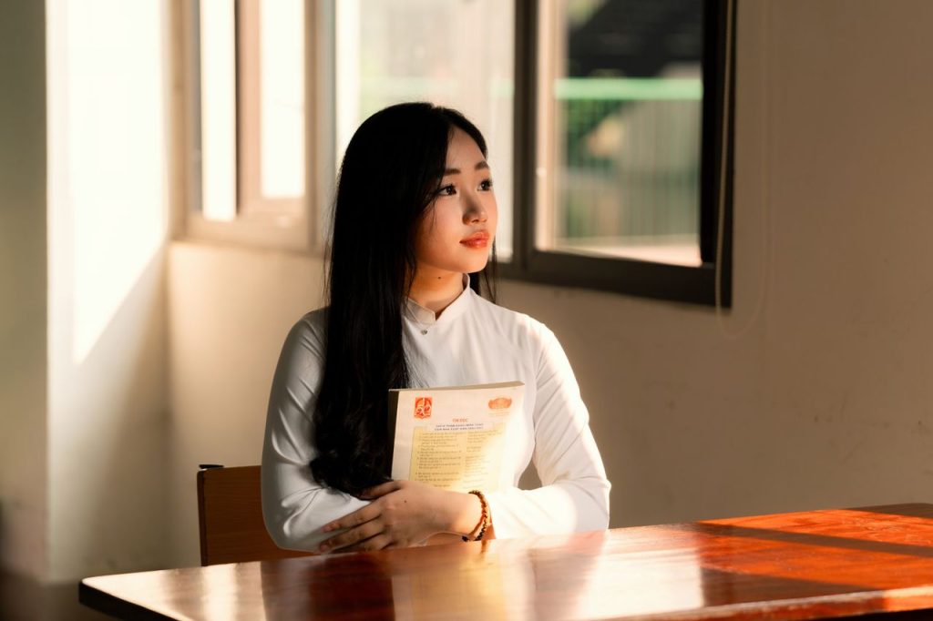 Beautiful female student in white Ao Dai sitting in a sunlit Vietnamese classroom.