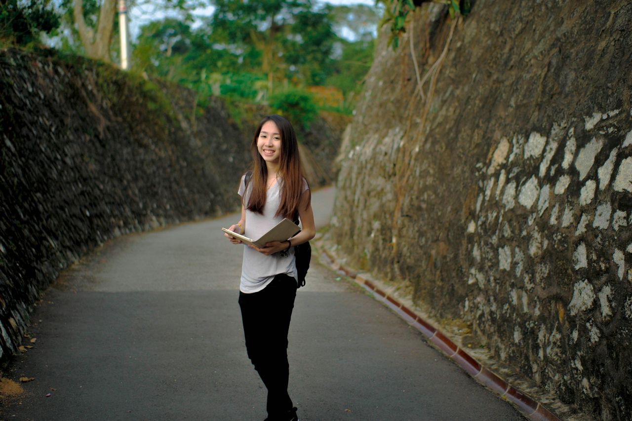 Mastering the First Impression: Your intriguing post title goes here Asian woman smiling while holding books on a stone pathway outdoors.